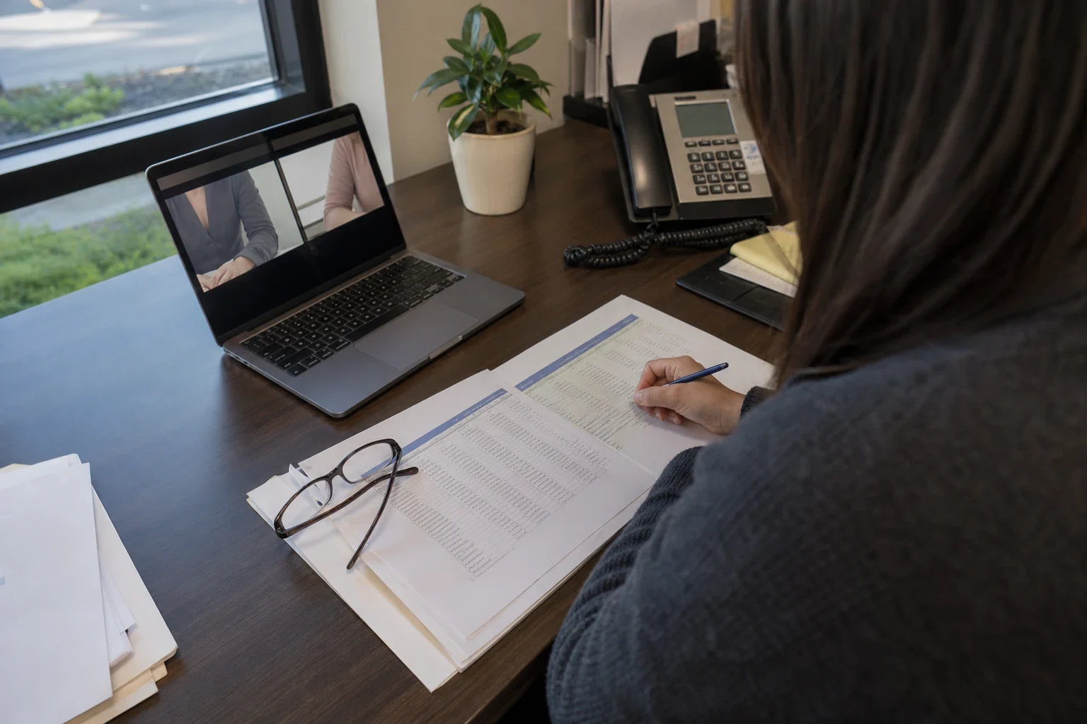 Agent seen from behind at an office desk, writing on a printed table of figures beside reading glasses, with a video call open on a laptop and a desk phone nearby