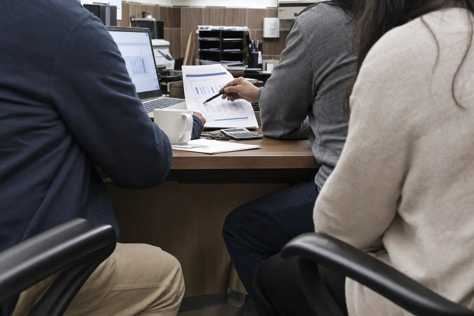 A man in a gray sweater points a pen at a printed bar chart while two people beside him follow along at a wooden office desk with a laptop and calculator