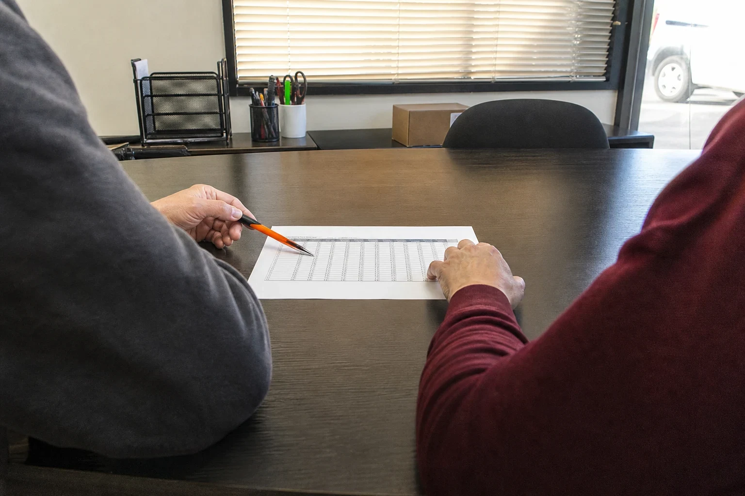 Two people at a dark office table, one pointing a pen at a printed table of figures while the other rests a hand on the page, with window blinds behind